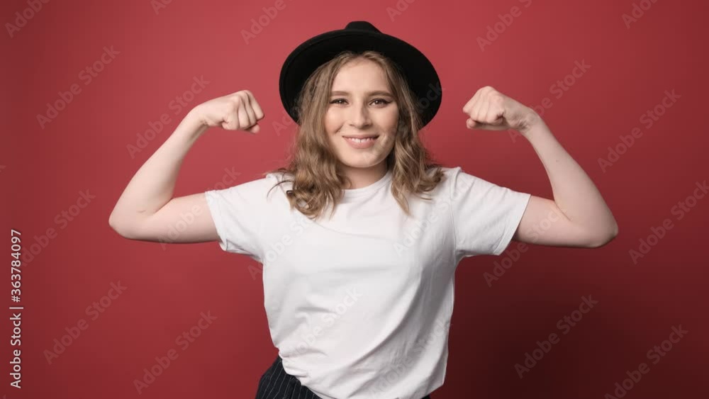 Attractive young woman raises arms and shows biceps, wears white t shirt and hat, isolated over red background Attractive young woman raises arms and shows biceps, wears white t shirt and hat, isolated over red background