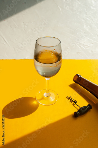 filled glass with water drops, dark glass wine bottle and corkscrew on a bright yellow background in natural light with sharp shadows, vertical image, summer bar menu photo