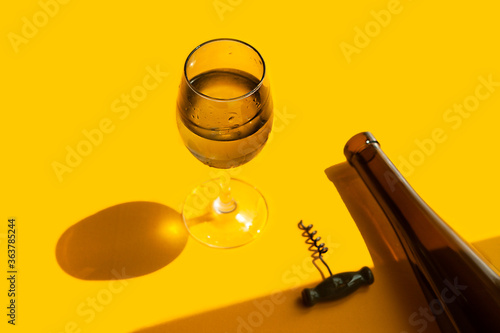 filled glass with water drops, dark glass wine bottle and corkscrew on a bright yellow background in natural light with sharp shadows, vertical image, summer bar menu
