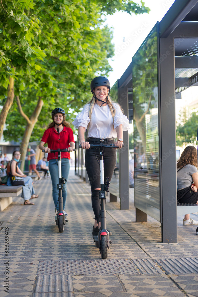 Trendy fashinable teenager girls riding public rental electric scooters in urban city environment. Eco-friendly modern public city transport. Trendy fashinable teenager girls riding public rental electric scooters in urban city environment. Eco-friendly modern public city transport.