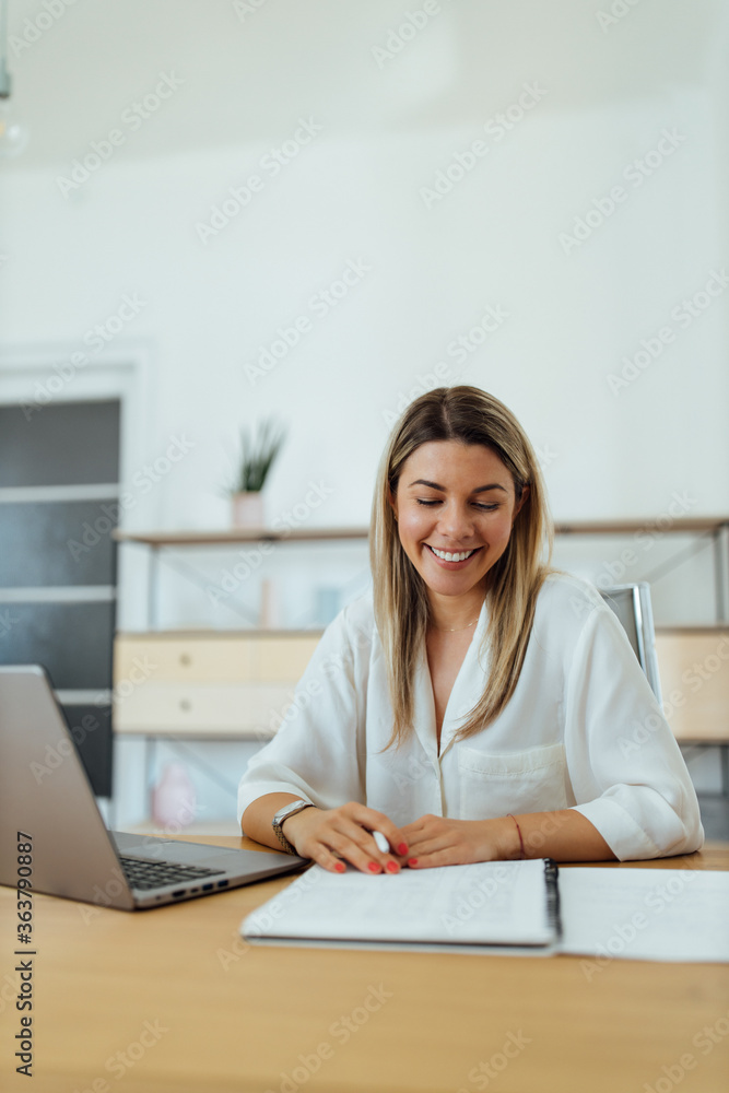 Portrait of a smiling woman sitting at desk with laptop, in the office, looking at notes. Portrait of a smiling woman sitting at desk with laptop, in the office, looking at notes.