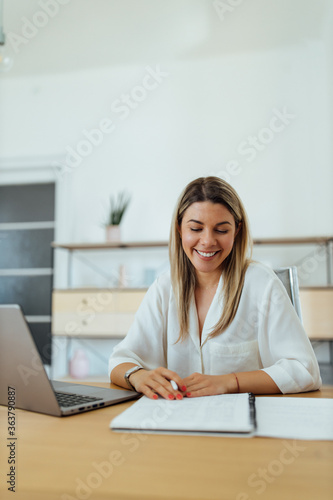 Portrait of a smiling woman sitting at desk with laptop, in the office, looking at notes.