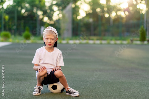 a blond boy in a cap in a sports uniform sits on a soccer ball on the football field, sports section. Training of children, children's leisure