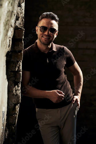 a stylish guy with glasses stands near a stone wall, wrapped himself and looks at the camera, smiles. strong and strict male gaze. brown T-shirt with collar and fashionable beige trousers. sunlight. 