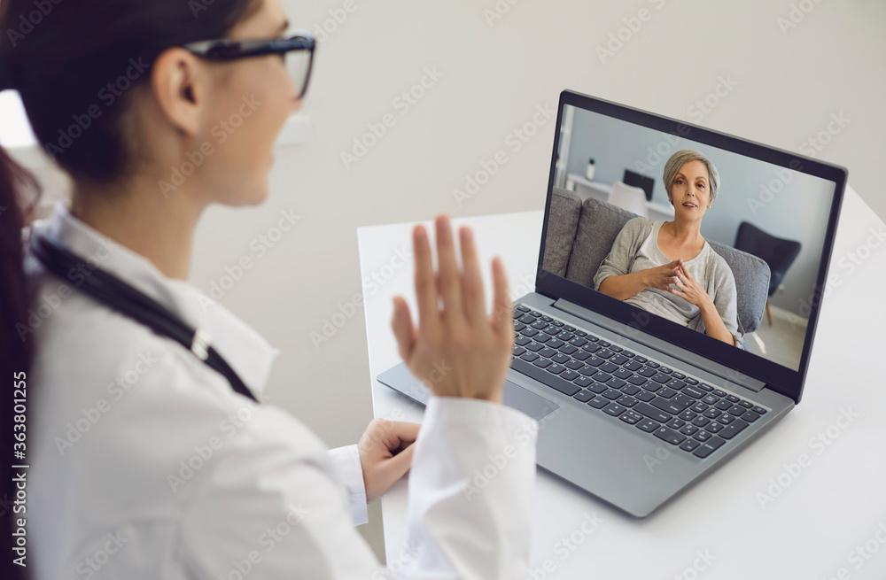 Girl doctor waves his hand consults an elderly patient woman using laptop sitting at a table in the office of the clinic. Girl doctor waves his hand consults an elderly patient woman using laptop sitting at a table in the office of the clinic.