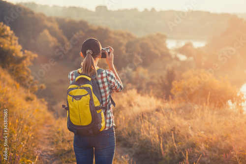 Fotografia, Obraz tourist girl with a backpack and a camera walks through a picturesque meadow and photographs the picturesque landscape