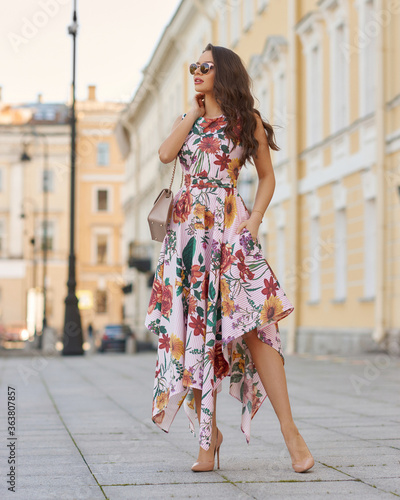 Young beautiful elegant tall slim woman with natural makeup and wavy brunete hair wearing colorful dress and sunglassses walking in the city on a summer day and holding handbag photo