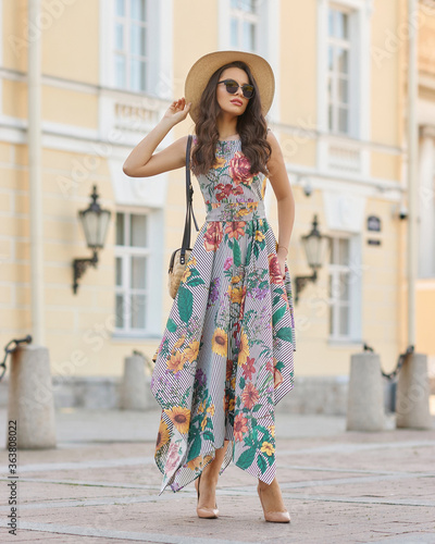 Young beautiful elegant tall slim woman with natural makeup and wavy brunete hair wearing colorful dress, salty hat and sunglassses walking in the city on a summer day and holding handbag