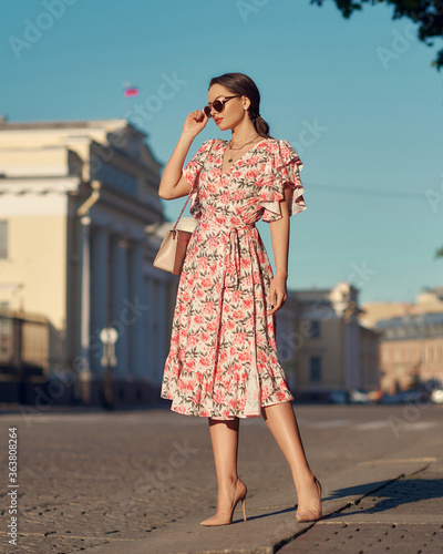 Pretty lady wearing wrap around dress. Beautiful woman with wavy brunette hair walking un the city. Elegant girl with long leegs in high heels. Fashionable female model in sunglasses holding handbag photo