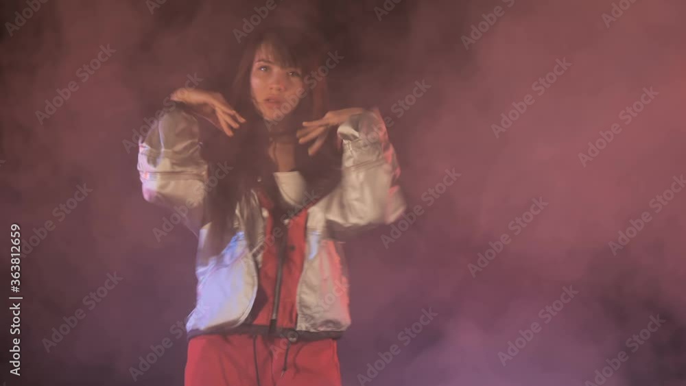 A young dancer with long hair in a sports silver sports jacket is dancing an active dance in clubs of pink smoke. She moves and sings along with close-up tunes. A young dancer with long hair in a sports silver sports jacket is dancing an active dance in clubs of pink smoke. She moves and sings along with close-up tunes.