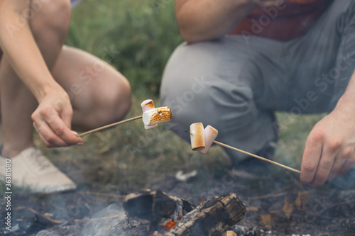 Guy and the girl are frying marshmallows on a campfire. Friends hold sticks with marshmallows over a bonfire. Horizontal orientation, selective focus.