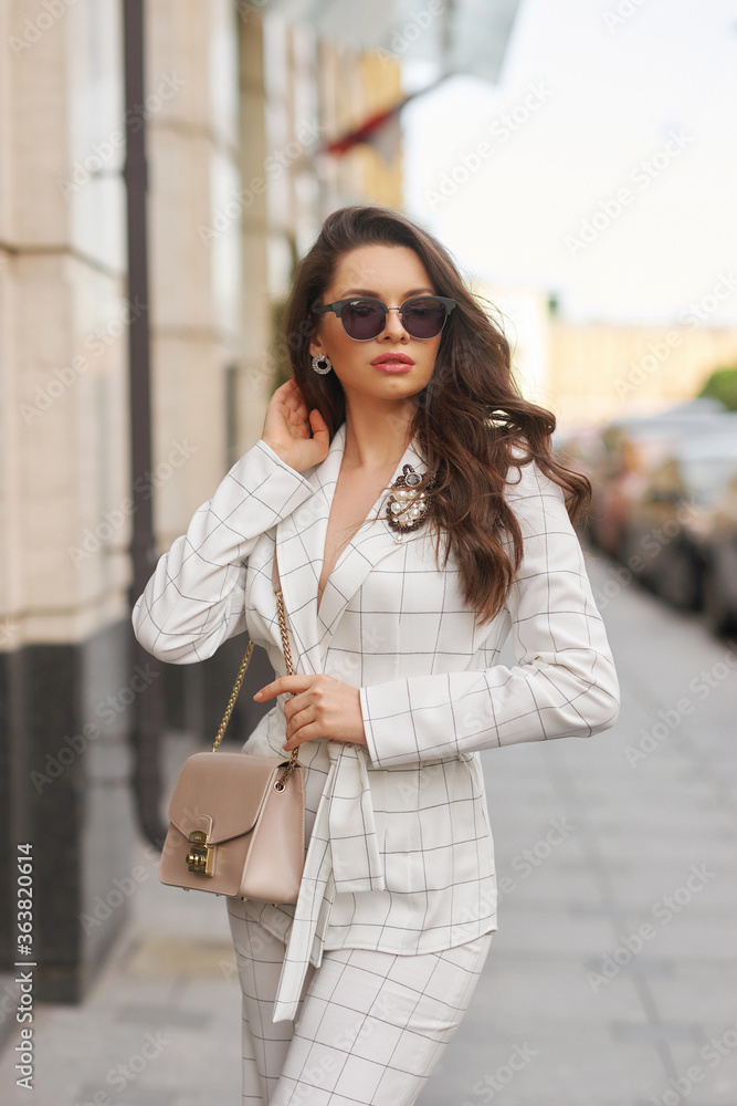 Young beautiful elegant woman in white casual costume walking at street on a summer day. Pretty lady wearing sunglasses and holding handbag. Businesswoman standing and posing in the city - obrazy, fototapety, plakaty 