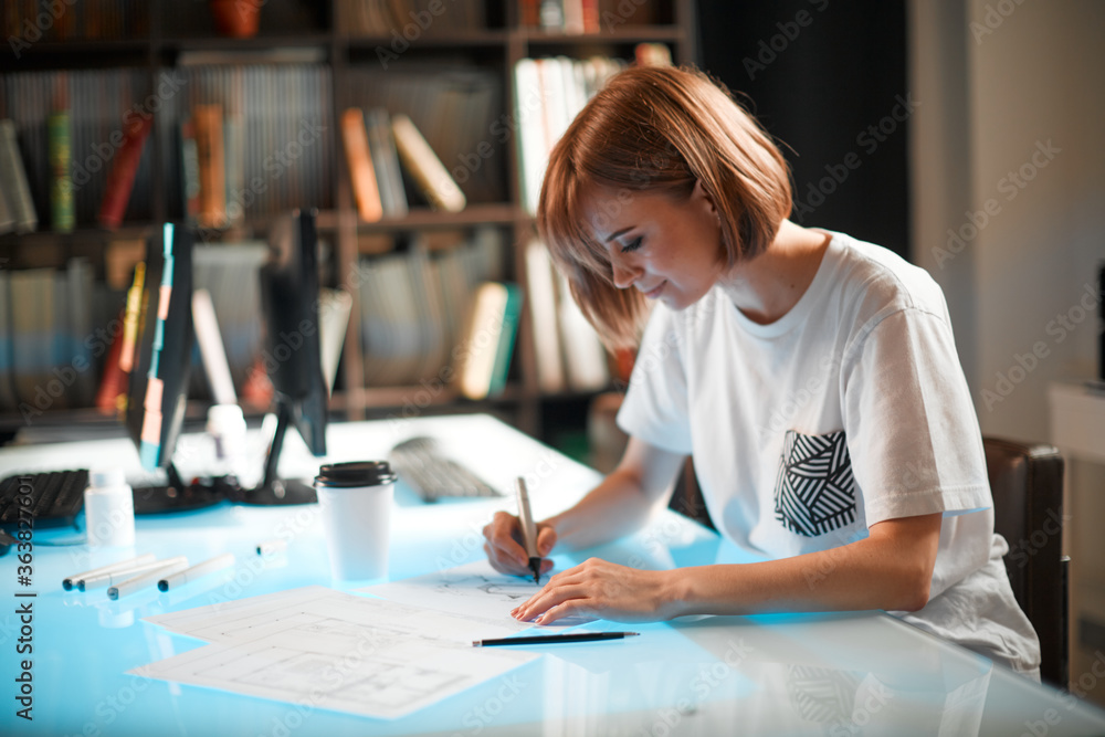 Young pretty designer is developing new project, workspace in creative company against the background of bookshelves Young pretty designer is developing new project, workspace in creative company against the background of bookshelves