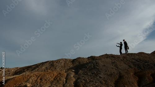 Two silhouettes of businessmen in the general plan move along the top of the hill against the sky. One of them holds a laptop in his hands and shows the second businessman with a briefcase the photo