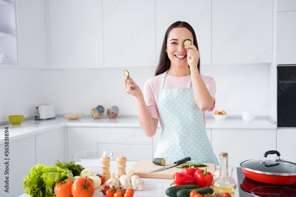 Photo of beautiful asian ethnicity housewife hold two fresh cucumber slices hiding one eye playful mood weekend cooking tasty breakfast salad stand kitchen indoors