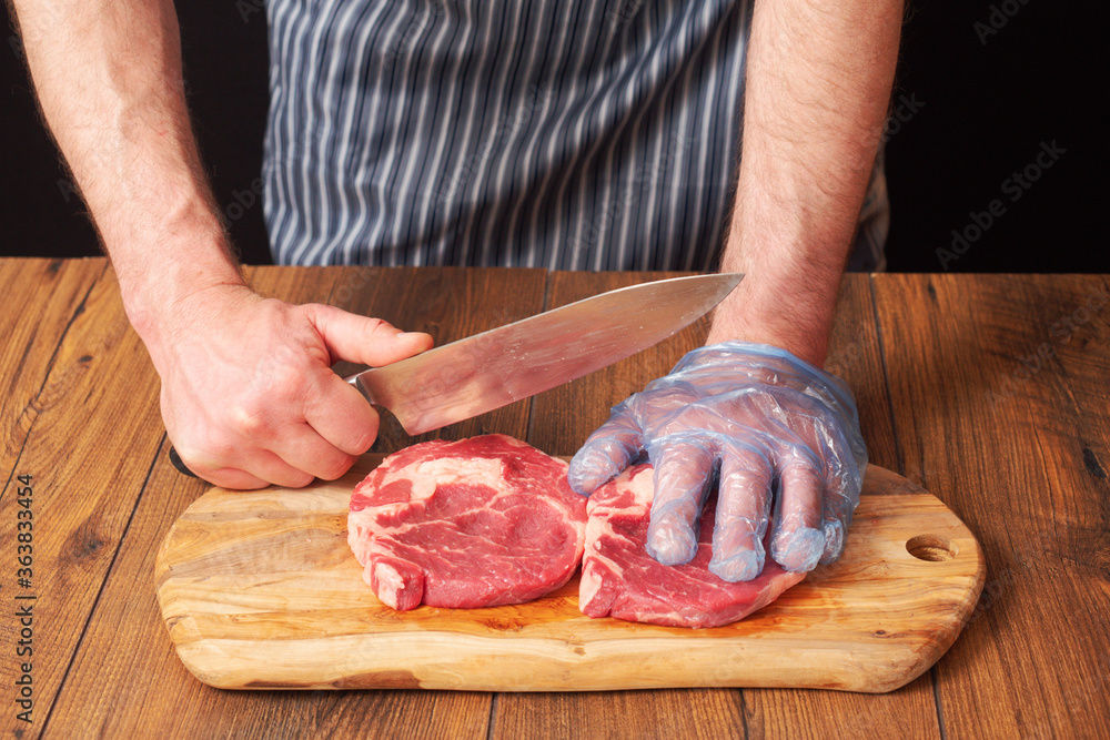 Butcher in black and white apron holding knife in his right hand, his left hand is on a freshly cut rib eye steak, Wooden board and table, black background. Meat industry.