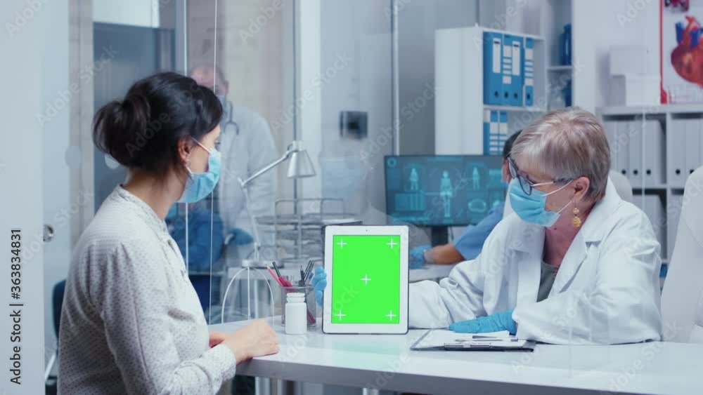 Senior doctor holding a green Screen tabled during COVID-19 health crisis, showing info to patient, wearing mask and protection gloves. Medical consultation in protective equipment concept shot of Senior doctor holding a green Screen tabled during COVID-19 health crisis, showing info to patient, wearing mask and protection gloves. Medical consultation in protective equipment concept shot of