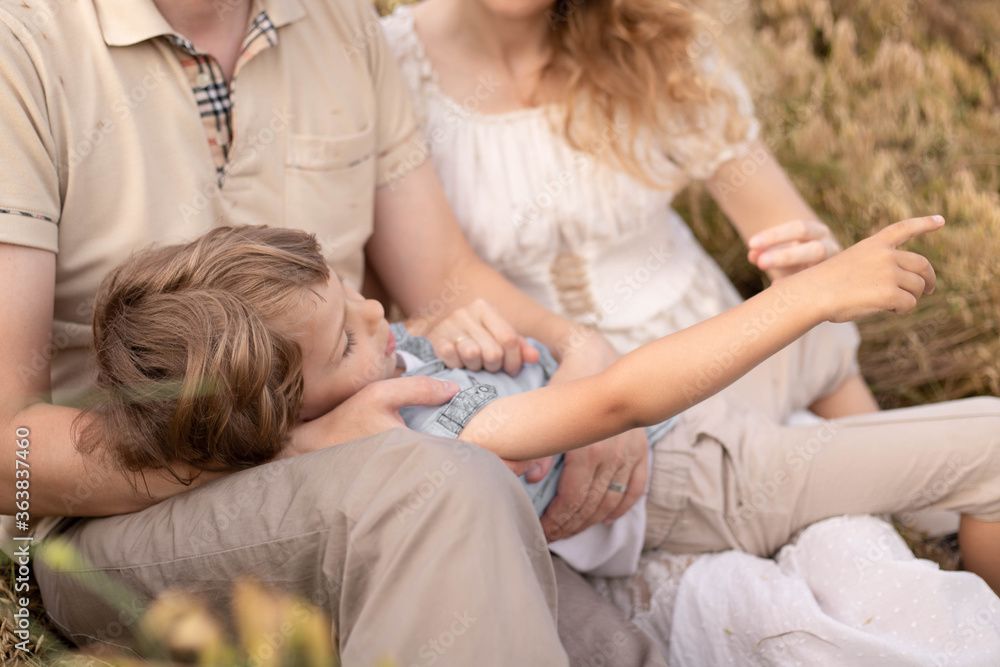 A little boy lies in the arms of his parents, a close-knit family, mom and dad, a happy child shows his parents a finger in the distance, people in the background of the field A little boy lies in the arms of his parents, a close-knit family, mom and dad, a happy child shows his parents a finger in the distance, people in the background of the field