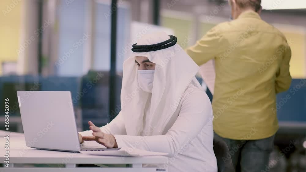 Workplace of an arab businessman during a pandemic. A man in Arabic clothes and a medical mask works in the office on a laptop. Work colleagues observe safety precautions. Workplace of an arab businessman during a pandemic. A man in Arabic clothes and a medical mask works in the office on a laptop. Work colleagues observe safety precautions.