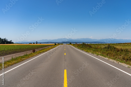 Wide open road. Symmetrical view down a two-lane road  flat landscape with fields either side and big blue sky and the sea at the end. Coupeville  Whidbey Island  Washington  USA.