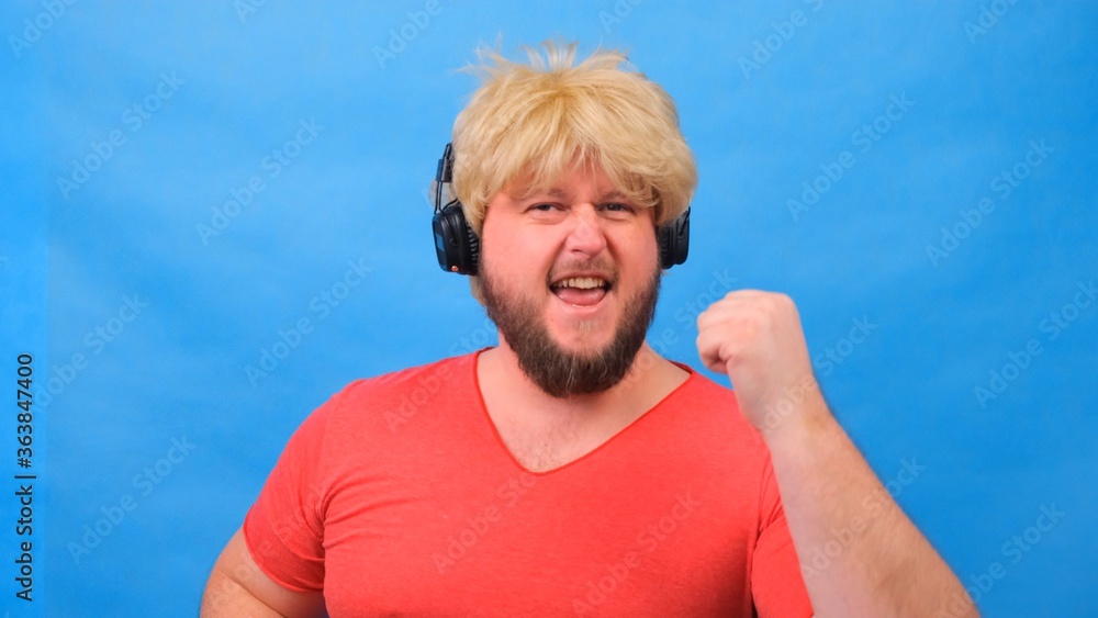 Naklejka premium freaky fat man in a wig and a pink T-shirt in his hands listens to music on headphones and dances on a blue background