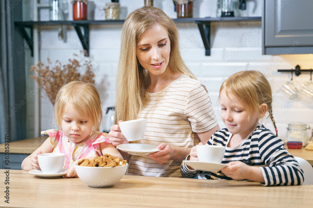 Happy family young mother and two daughters drinking tea or milk with freshly baked homemade cookies in the kitchen Happy family young mother and two daughters drinking tea or milk with freshly baked homemade cookies in the kitchen