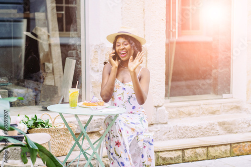 Portrait of a relaxed young african american woman drinking lemonade, eating dessert, sitting at cozy outdoor cafe with green plant talking on cell phone.. Sun glare effect.