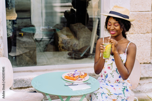 Young smiling african american girl drinking tasty sweet cocktail , amazing relaxing day, tasty lemonade, elegant dress and straw hat, outdoor cafe, summer mood.