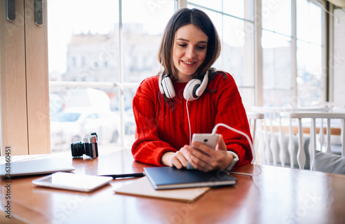 Cheerful female teenager with headphones connecting to cellular surfing and messaging in online chat using 4G internet.Internet user searching music on websites for loading on telephone in cafe