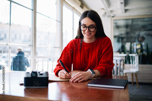 Smiling positive female student journalist sitting at table and writing essay exited with idea, successful hipster girl in eyeglasses enjoying time at university campus while completing homework