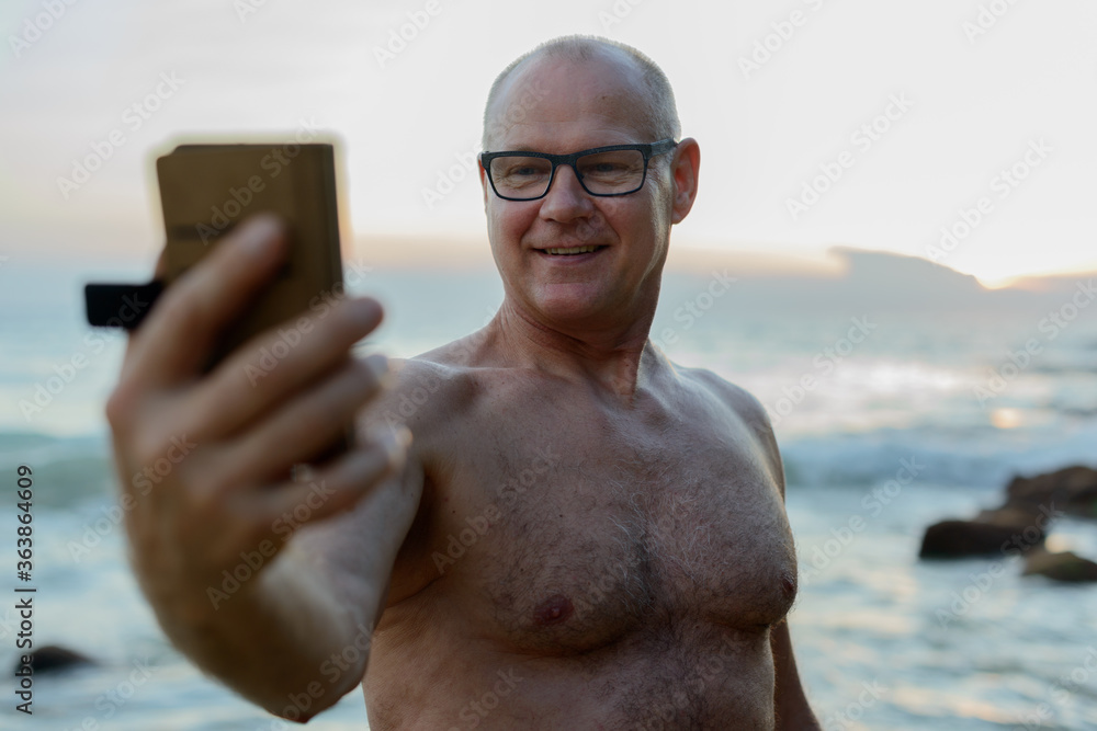Happy mature shirtless tourist man using phone at the beach