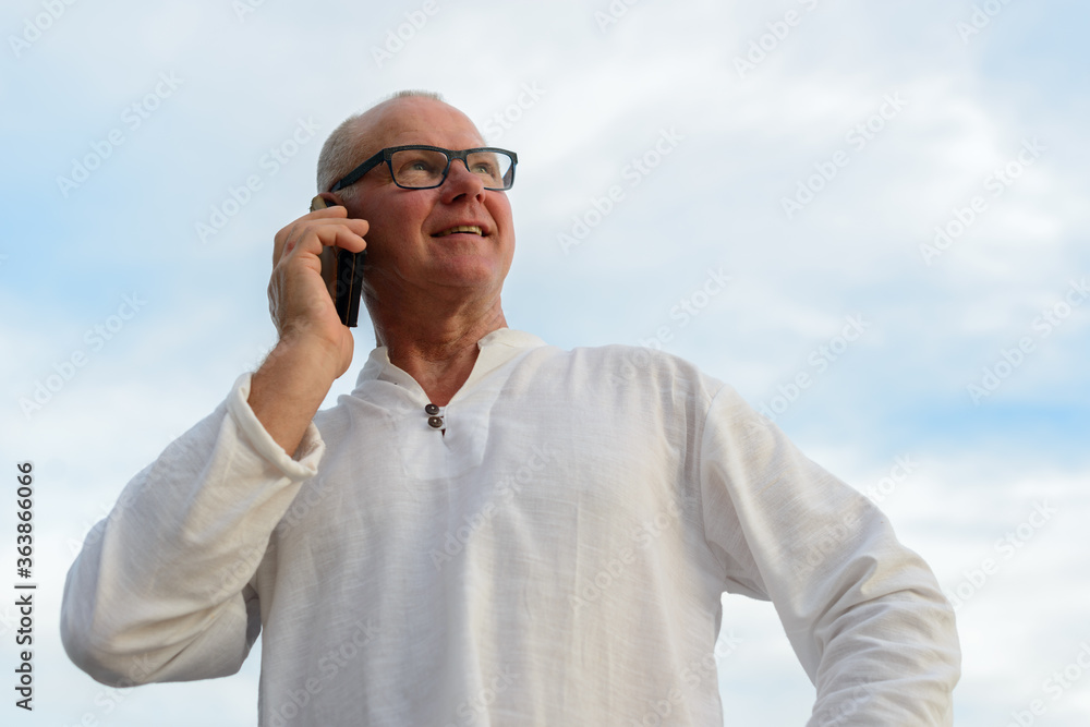 Happy mature handsome tourist man talking on the phone against view of the beach outdoors