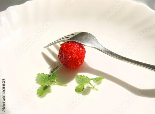  single raw ripe strawberry on a white plate with mint leaves and a fork 