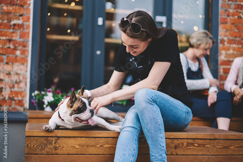 Happy hipster girl in casual apparel feeling excited of spending leisure together with favourite home pet, positive woman resting with french bulldog enjoying friendly time with animal on bench
