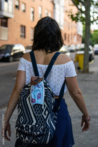 Photo of a young and attractive woman wearing a backpack with a disposable face mask attached to it with lipstick on it walking down the street