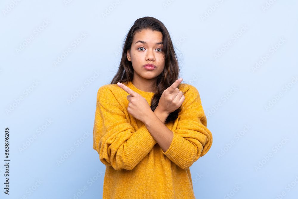 Young Colombian girl with sweater over isolated blue background pointing to the laterals having doubts Young Colombian girl with sweater over isolated blue background pointing to the laterals having doubts