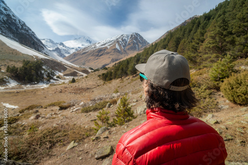 A long-haired young man in a red down jacket and grey cap in sunglasses stands with his back to the camera and looks at a mountain gorge with snow-capped peaks in the distance