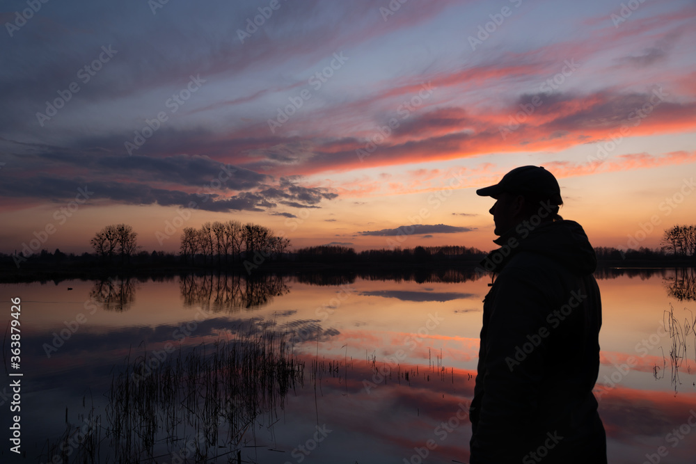 The silhouette of a man on the shore of a lake and sky by sunset