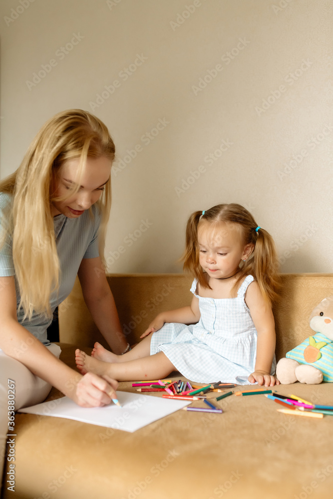 Young beautiful mother blonde and her daughter 2 years old draw with pencils on the sofa in the living room at home Young beautiful mother blonde and her daughter 2 years old draw with pencils on the sofa in the living room at home