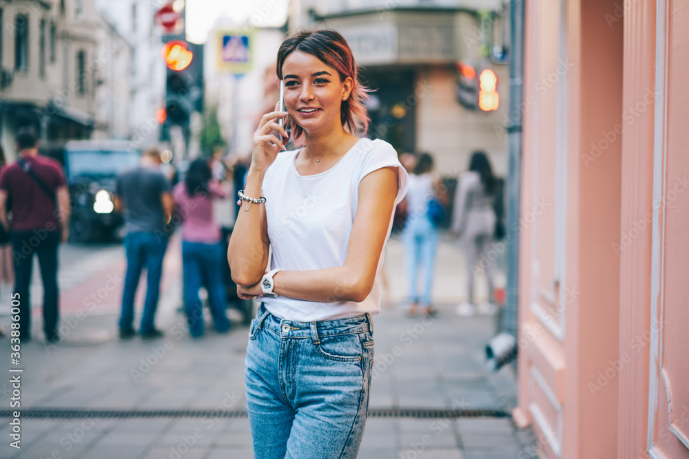 Pretty hipster girl with short pink hair talking on modern cellular standing on street.Positive female dressed in t-shirt with mock up area for advertising having friendly conversation on mobile phone