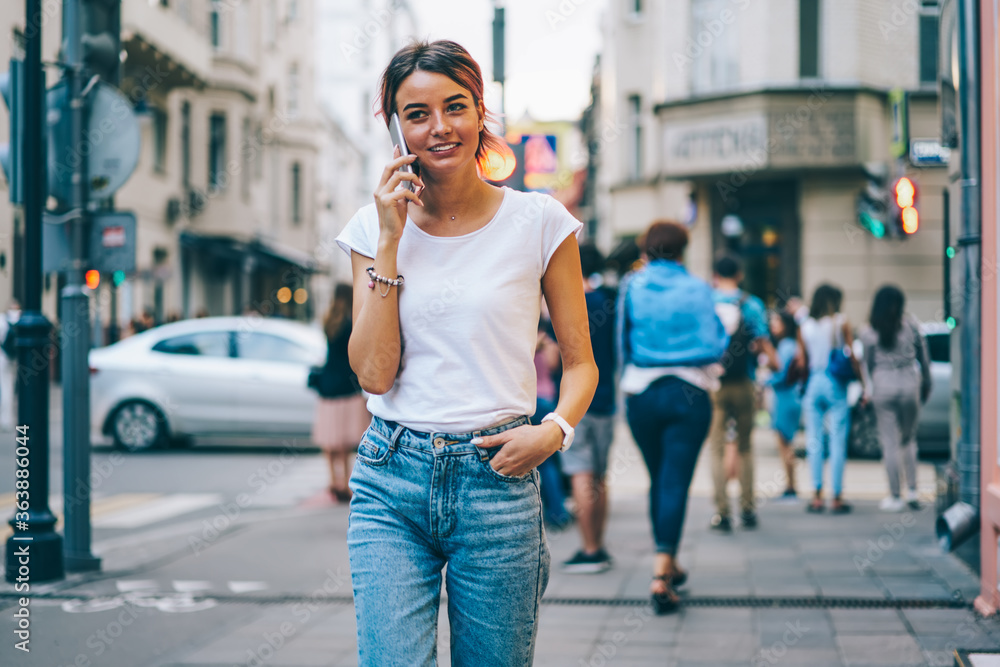 Stylish cheerful young woman dressed in white t-shirt with mock up area for text message strolling in street of modern city and talking with friend on telephone.Colorful promotional background