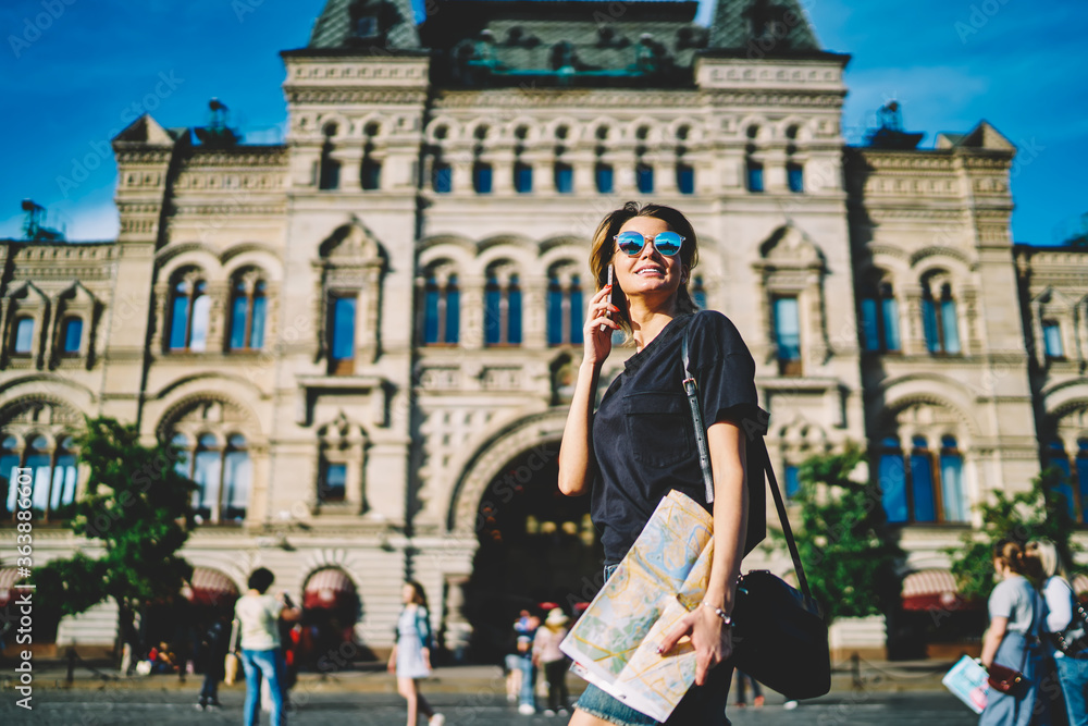 Young smiling female traveler having mobile conversation with friends via useful smartphone application while strolling at urban setting.Happy blogger in sunglasses talking on cellular outdoors