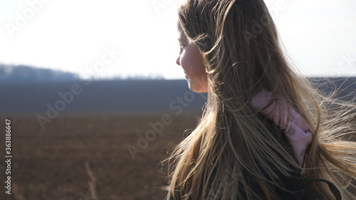Small smiling girl with blowing blonde hair walks on the road near plowed field. Happy kid with backpack moves on way to school. Female child goes along a driveway near ploughed meadow. Dolly shot