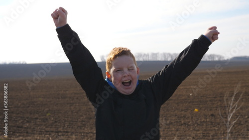 Little red-haired boy with freckles looks into camera and raises hands rejoicing achievement standing on ploughed meadow. Portrait of happy male child with positive emotion on his face at plowed field