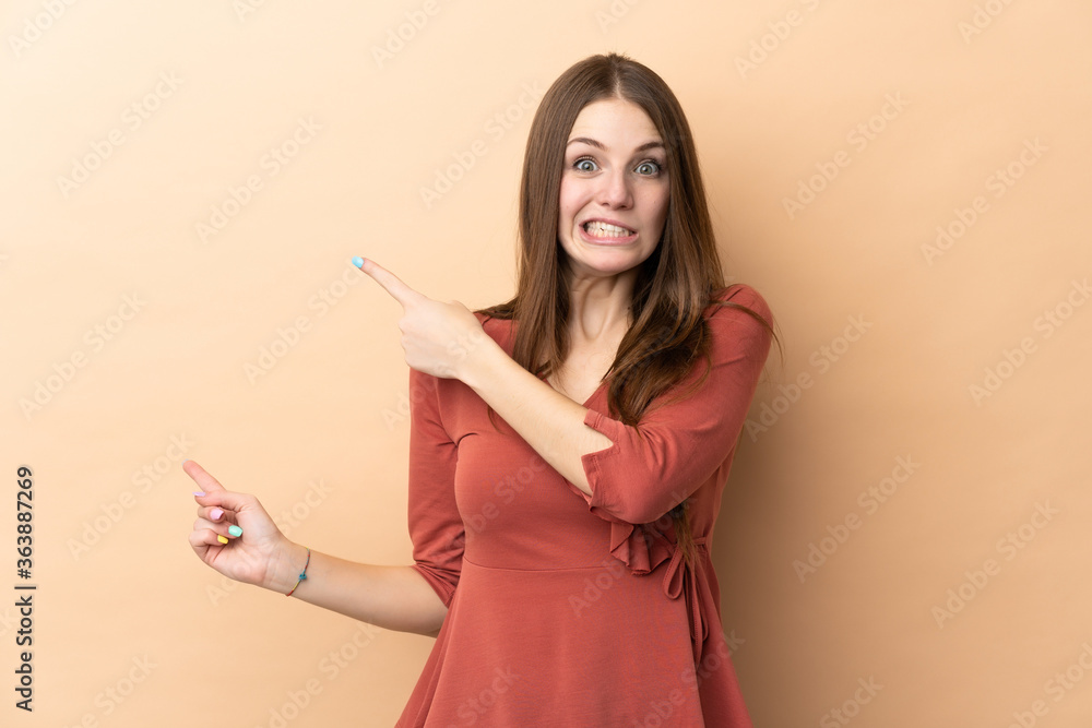 Young caucasian woman isolated on beige background frightened and pointing to the side Young caucasian woman isolated on beige background frightened and pointing to the side