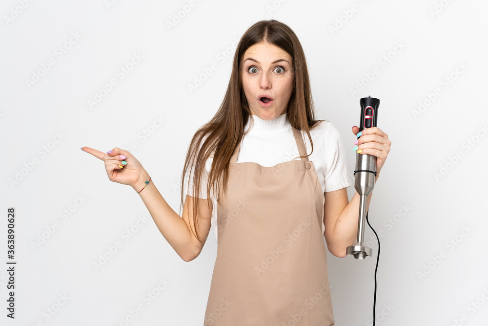 Young woman using hand blender isolated on white background surprised and pointing finger to the side Young woman using hand blender isolated on white background surprised and pointing finger to the side