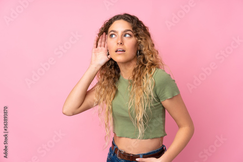 Young blonde woman with curly hair isolated on pink background listening to something by putting hand on the ear © luismolinero