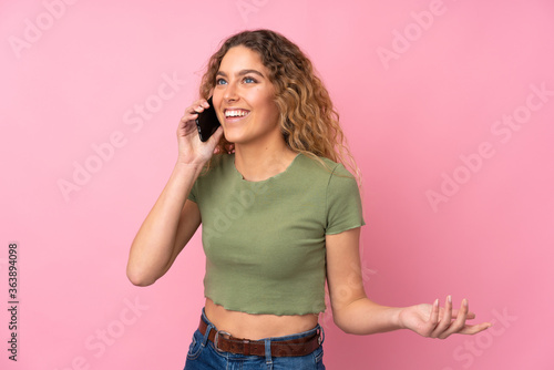 Young blonde woman with curly hair isolated on pink background keeping a conversation with the mobile phone with someone