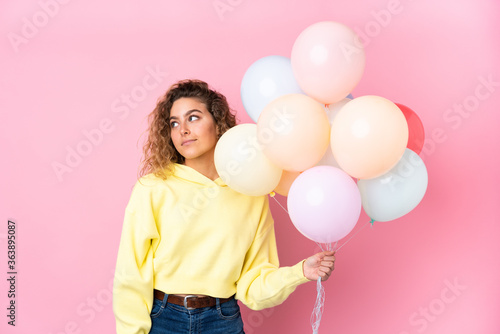 Young blonde woman with curly hair catching many balloons isolated on pink background laughing and looking up