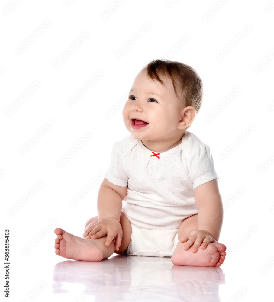 Adorable little baby infant toddler in white cotton bodysuit sits on floor, laughing, touching legs and looking aside Adorable little baby infant toddler in white cotton bodysuit sits on floor, laughing, touching legs and looking aside